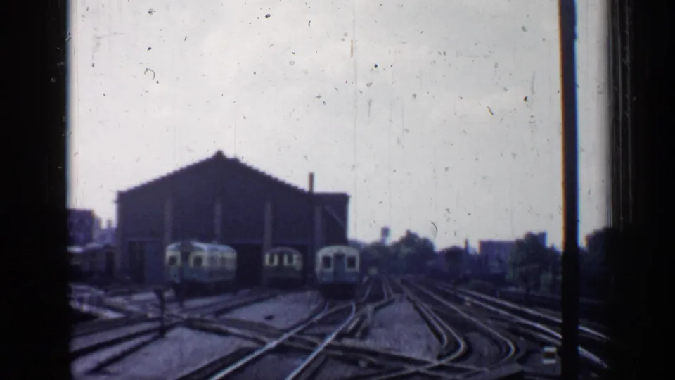 1958 Chicago rail-yard view from a moving train, with intersecting tracks, pale train cars, a large shed, and distant buildings.