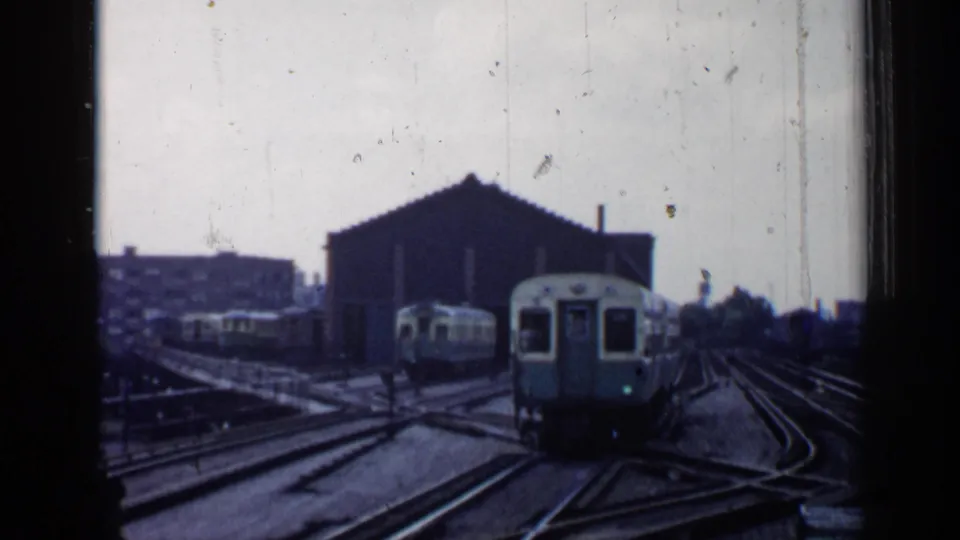 A light-colored train moves through the 1958 Chicago rail yard as tracks converge near a dark shed and city buildings.