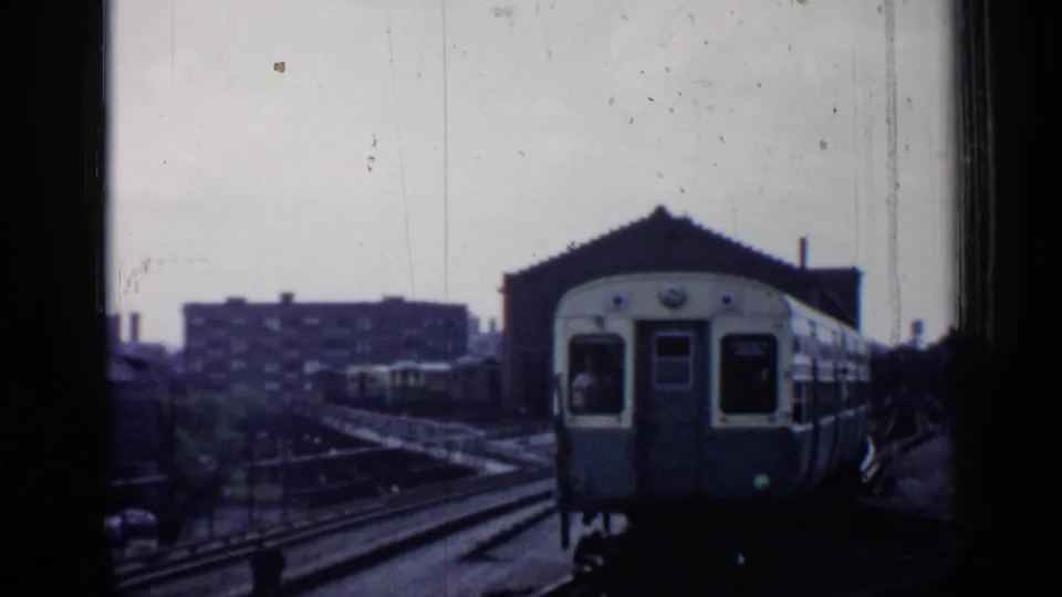 The front of a pale train comes close to the passenger viewpoint in a Chicago rail yard, with buildings and tracks behind it.