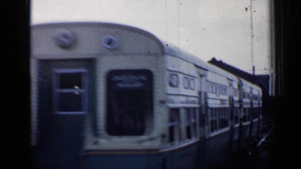 A pale train passes close beside the moving camera in the 1958 Chicago rail yard, producing strong motion blur across the car side.