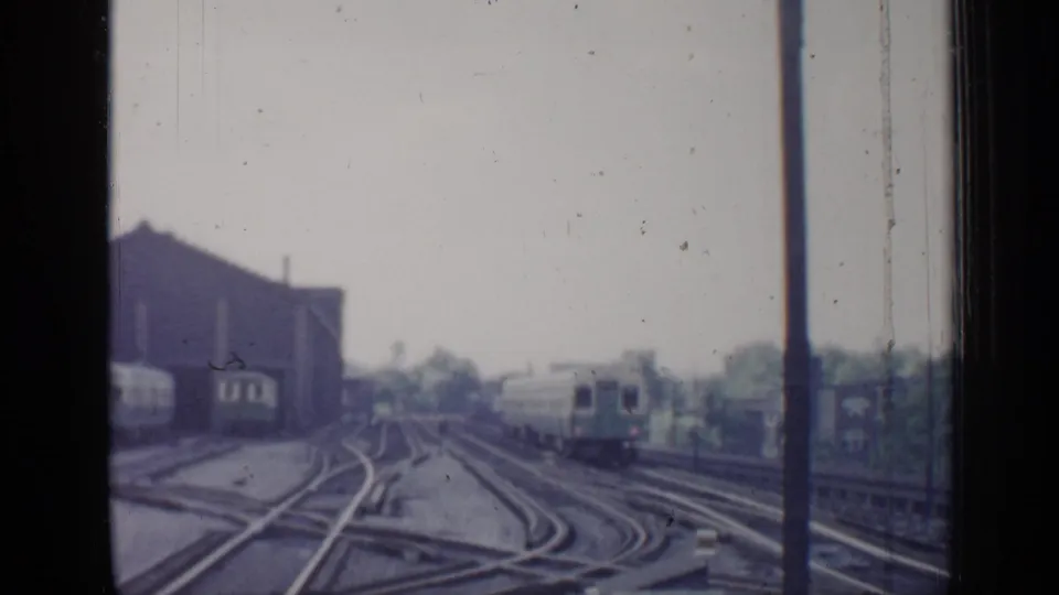 1958 Chicago rail scene showing a distant passenger train, multiple track switches, station-side structures, and soft 8mm exposure.