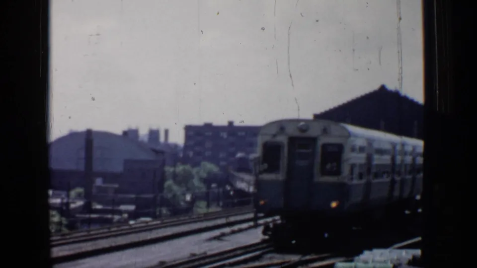 A two-car commuter train approaches through a Chicago rail yard, framed by window edges and mid-century industrial buildings.