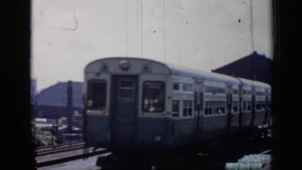 The front car of a 1958 Chicago commuter train fills the frame as it moves past rail yard buildings and tracks.