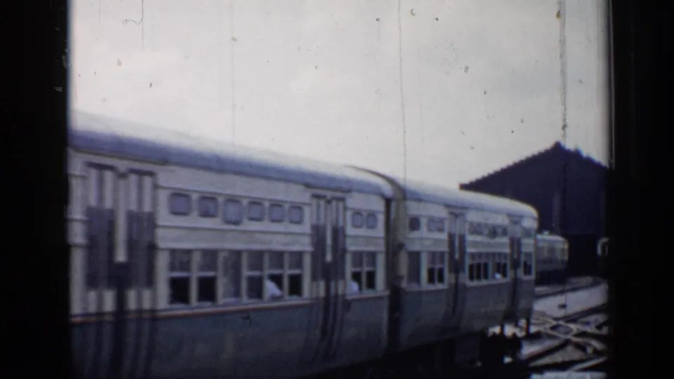 Side view of a two-car commuter train moving through a Chicago rail yard, with switches and buildings visible behind it.
