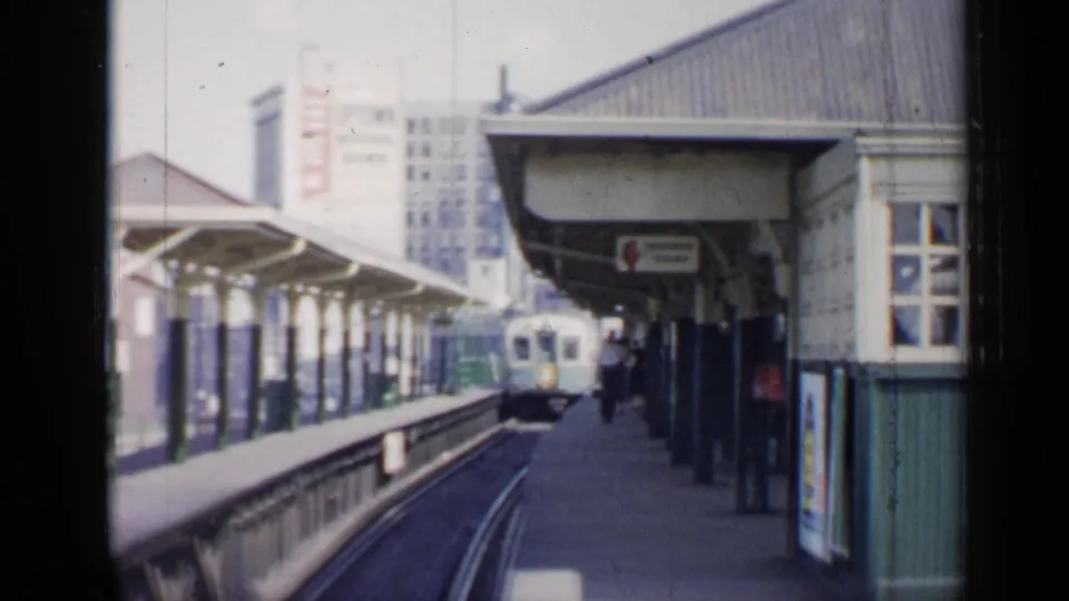 1958 Chicago elevated train approaching a covered platform, with tracks, columns, signs, and city buildings visible.