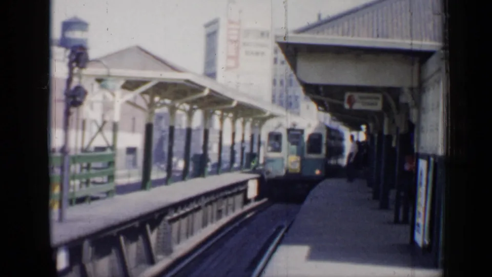 The train reaches the elevated Chicago platform, with waiting passengers, station posts, posters, and downtown buildings visible through soft 8mm grain.