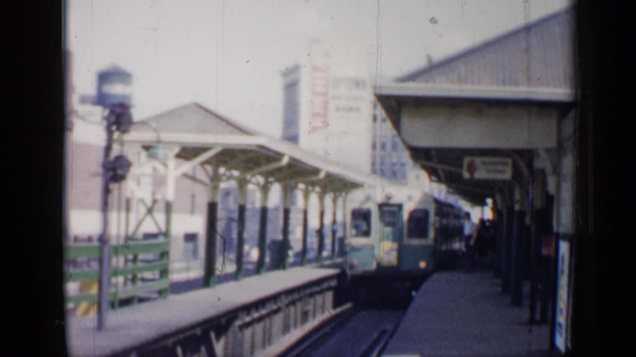 Green-and-white rail car approaching a covered station platform with city buildings behind.