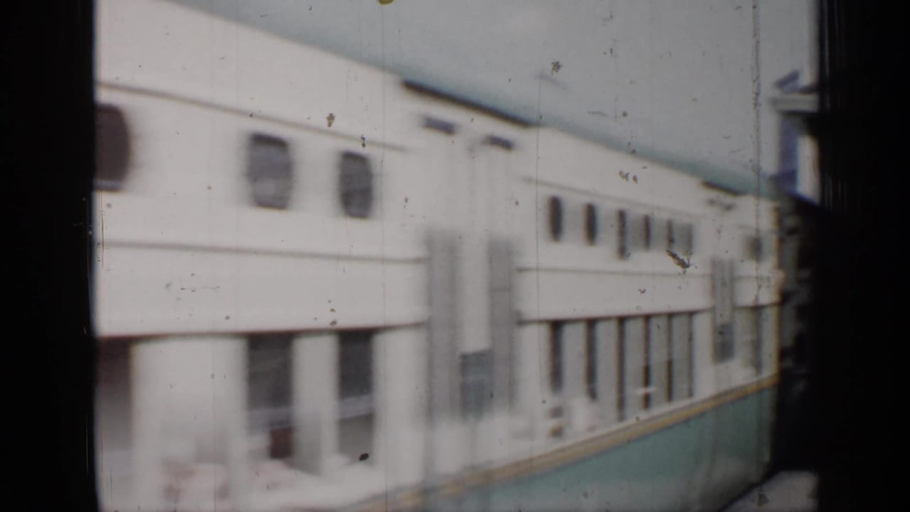 White and teal passenger train passing a station platform with dark film-frame edges.