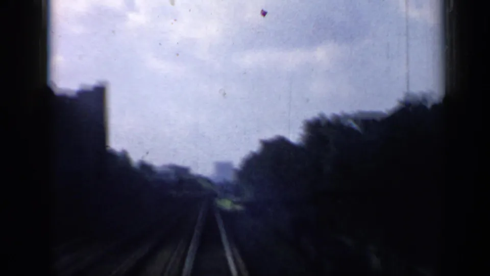 Soft 8mm view from a moving train in Chicago, looking down tracks past trees toward faint distant buildings.