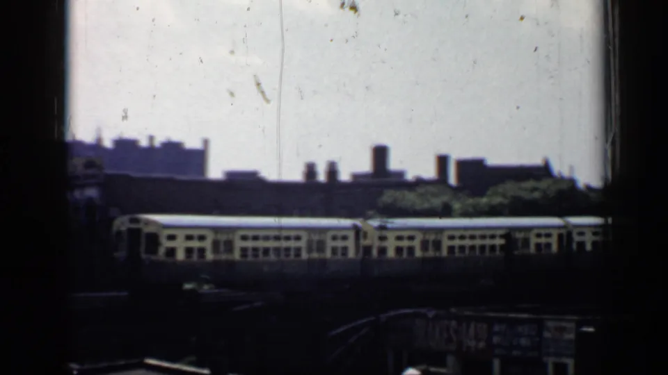 1958 Chicago home-movie frame with a train car passing on elevated tracks, seen through heavy dark framing above street-level signage.