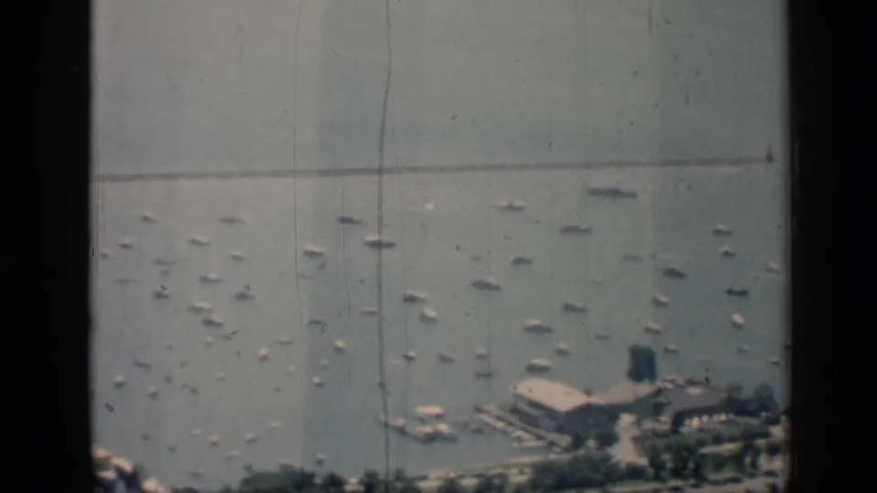 1958 Chicago harbor footage showing scattered boats, shoreline piers, a low building, trees, and dark vertical window framing.