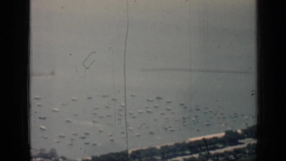 1958 Chicago harbor frame showing many small anchored boats, shoreline greenery, a long pier or breakwater, and pronounced 8mm film scratches.