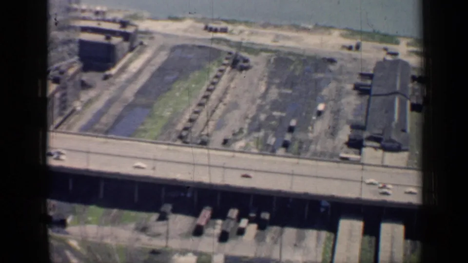 A high-angle Chicago waterfront frame shows a highway with cars, industrial shoreline lots, water channels, and dock structures in 1958 home-movie footage.
