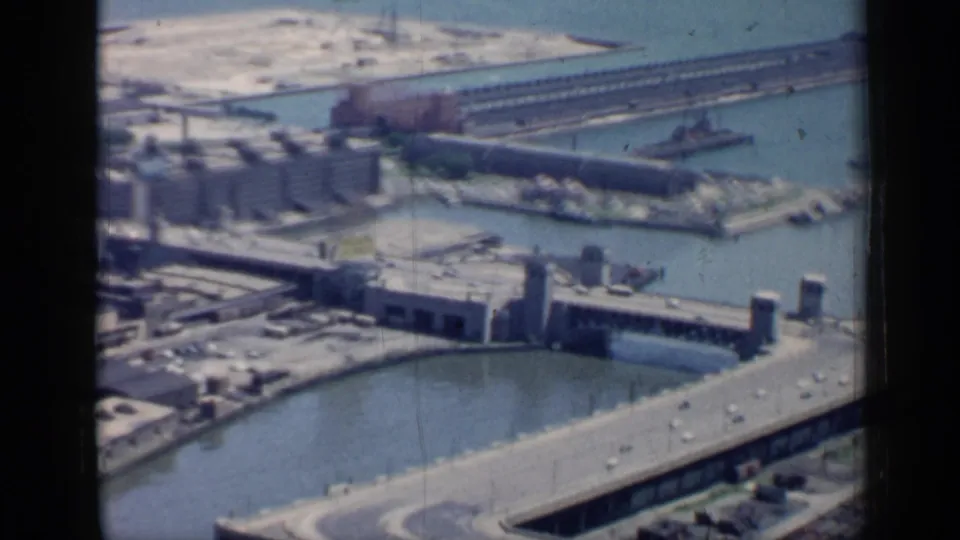 1958 Chicago aerial frame showing industrial waterfront buildings, canal water, bridge or lock structures, roadway traffic, and lakefront piers.
