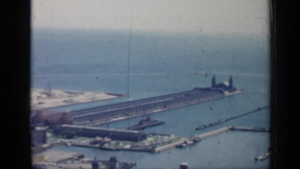 1958 Chicago aerial frame with a long waterfront pier, open lake water, small boats, shoreline, and industrial structures near the water.