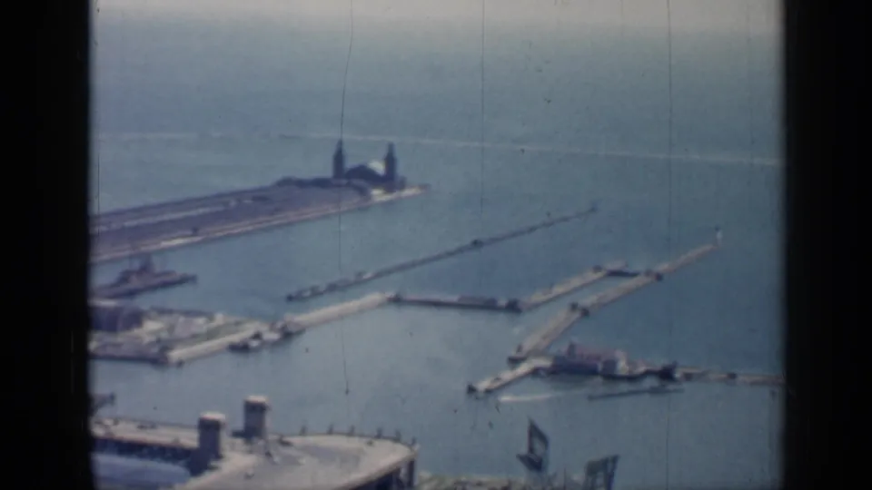 1958 Chicago aerial frame looking across harbor breakwaters, boats, open Lake Michigan water, pier-end structures, and a waterfront roofline below.