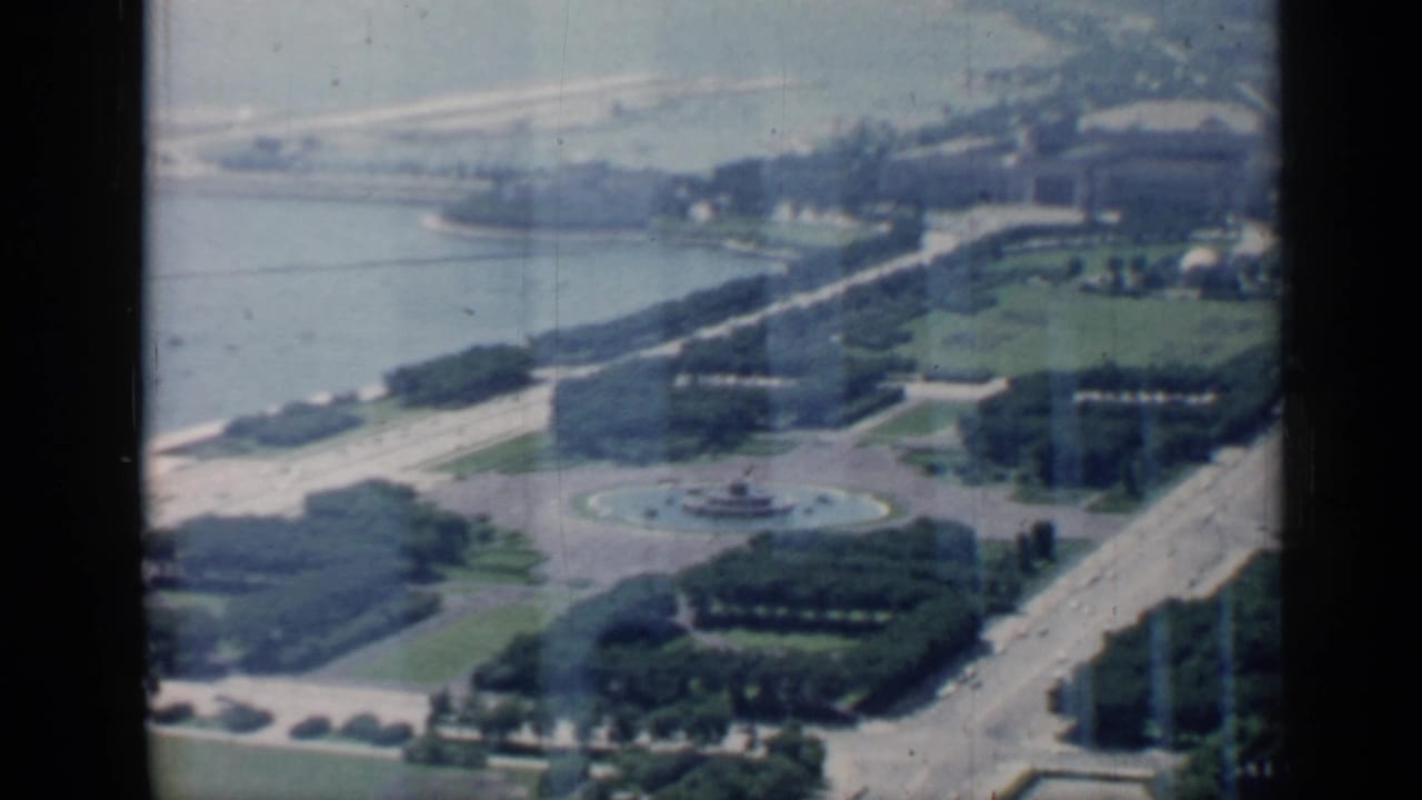 Aerial view of a lakefront park, roads, trees, and a circular fountain below.