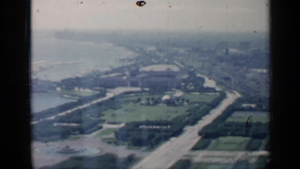 1958 Chicago aerial frame showing lakefront roads, green park space, shoreline, a circular fountain, and distant city forms.