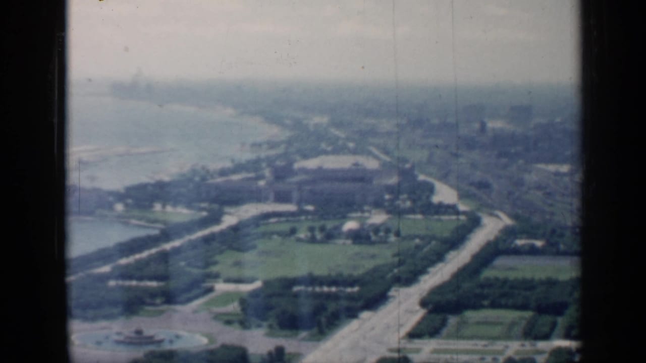 Aerial view over lakefront parkland, roads, and distant urban blocks framed by dark aircraft-window edges.