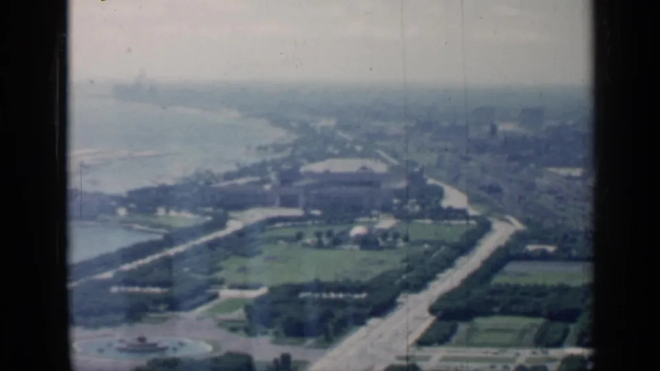 1958 Chicago aerial view showing lakefront land, curving roads, green park areas, and distant city blocks through a dark window frame.