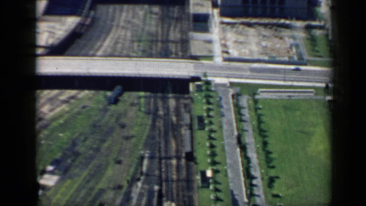 1958 Chicago aerial frame with a road bridge over multiple rail tracks, green verge, tree-lined road, and nearby urban structures.