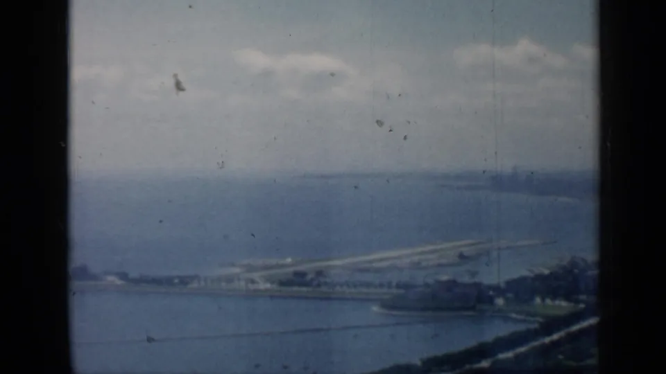 Aerial 8mm view over the Chicago lakefront, with water in the foreground, a long shoreline structure, and muted clouds above.