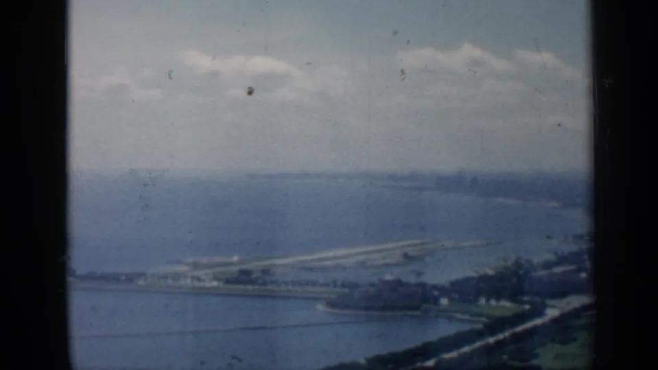 1958 Chicago lakefront aerial view with shoreline greenery, a pier or breakwater, open water, and faint city forms in the distance.