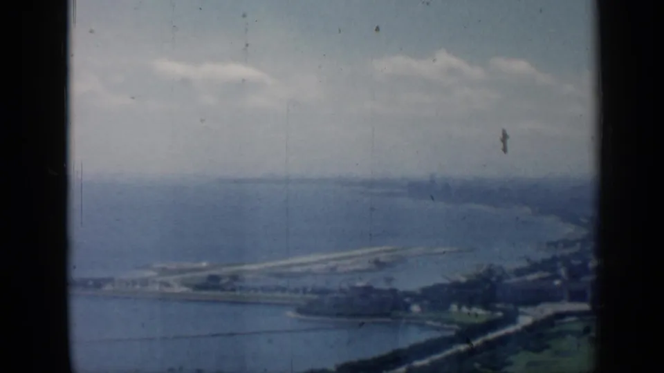 1958 Chicago-area aerial view toward Lake Michigan shoreline, softened by haze and framed by dark 8mm film edges.