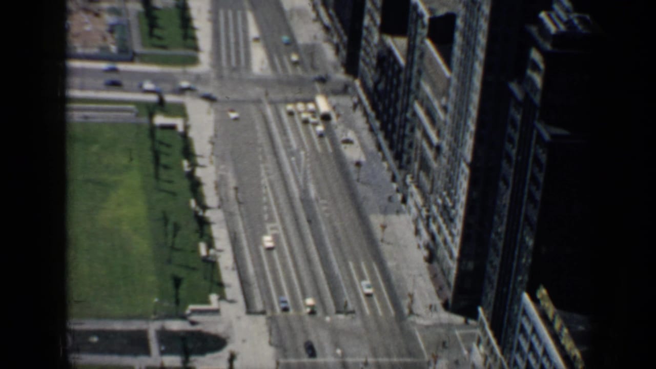 High-angle view of a broad city avenue beside green space and tall buildings.