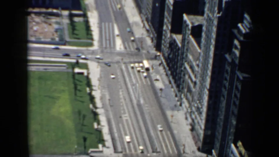 1958 Chicago aerial street view showing green lawn, a broad intersection, small cars, sidewalks, and shadowed high-rise facades.