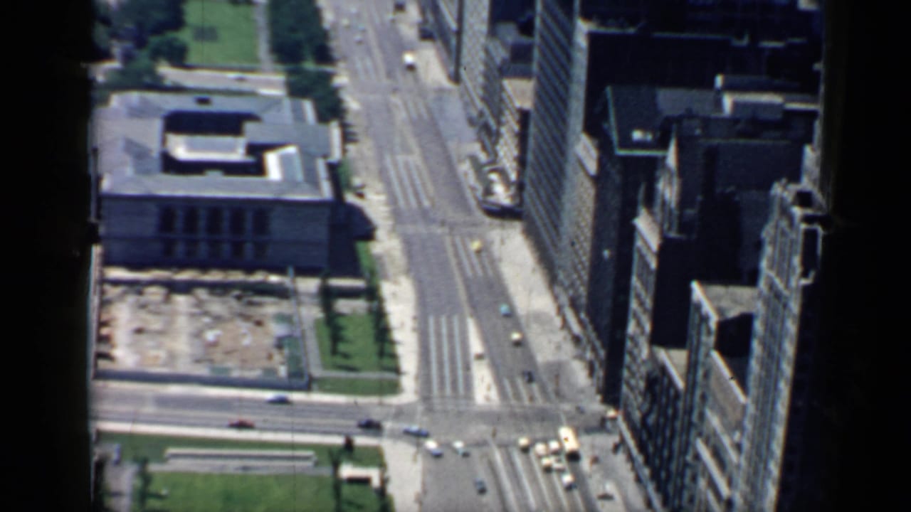 High-angle downtown street view with tall buildings, cars, lawns, and a flat-roofed civic-style structure.