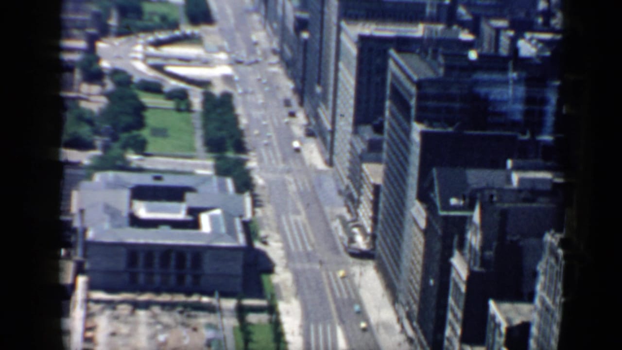Aerial city view with a broad road, high-rise facades, green park areas, and a curved paved feature.