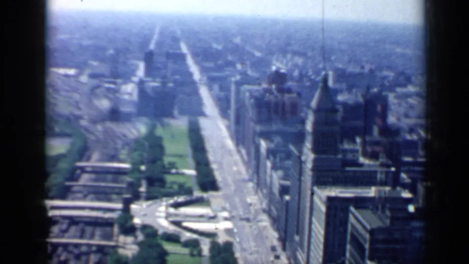 Wide 1958 Chicago aerial view looking along a straight avenue with park greenery, downtown towers, distant haze, and dark 8mm frame edges.