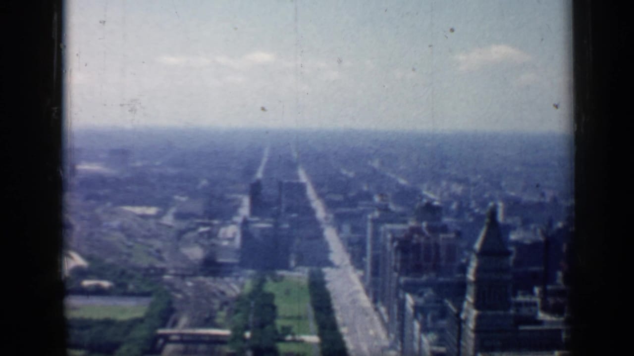 Aerial city view with a long straight street, high-rises, and park areas under a pale sky.