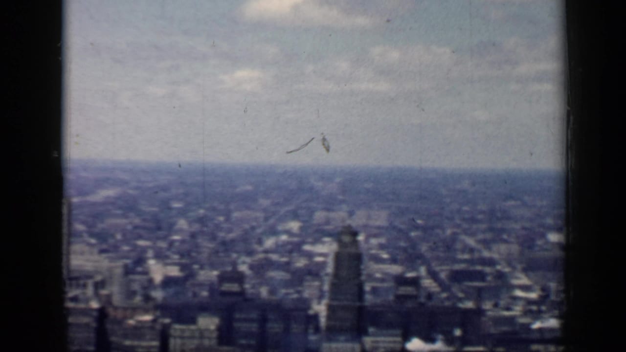 Wide city view with distant buildings, street grid, sky, and a dark vertical window bar.