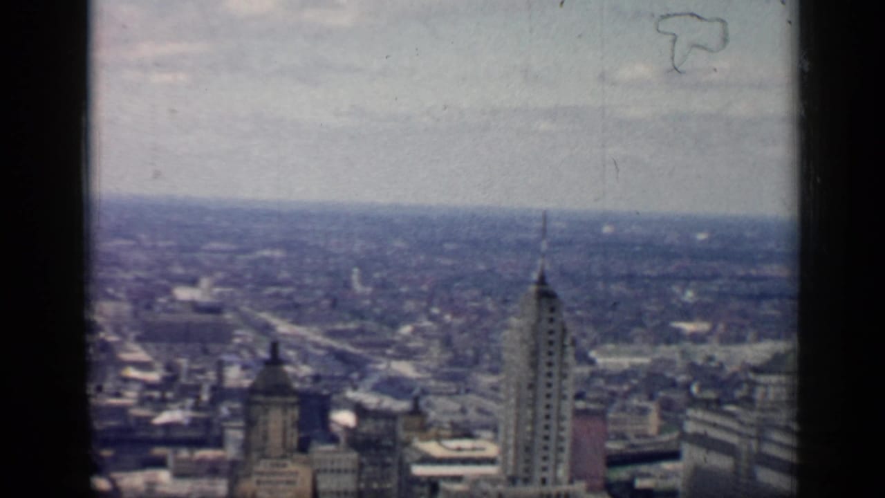 1958 8mm high-angle view over downtown Chicago, with tall skyscraper tops, a hazy city horizon, and dark film-frame edges.
