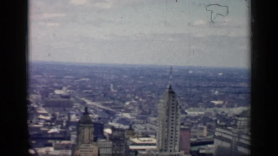 1958 8mm high-angle view over downtown Chicago, with tall skyscraper tops, a hazy city horizon, and dark film-frame edges.
