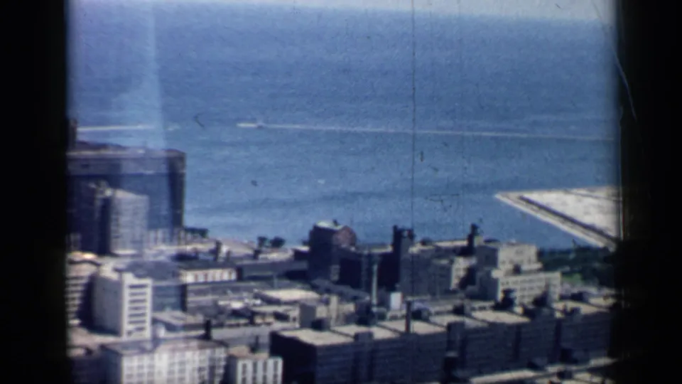 1958 Chicago home-movie frame with mid-century buildings in the foreground and Lake Michigan with shoreline infrastructure beyond.