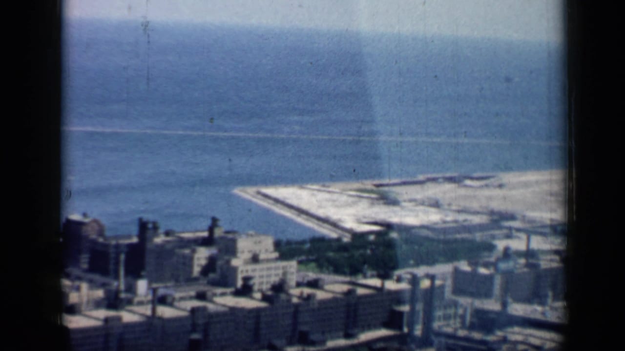 1958 Chicago aerial home-movie frame showing shoreline buildings, blue lake water, and a pale pier or breakwater along the coast.