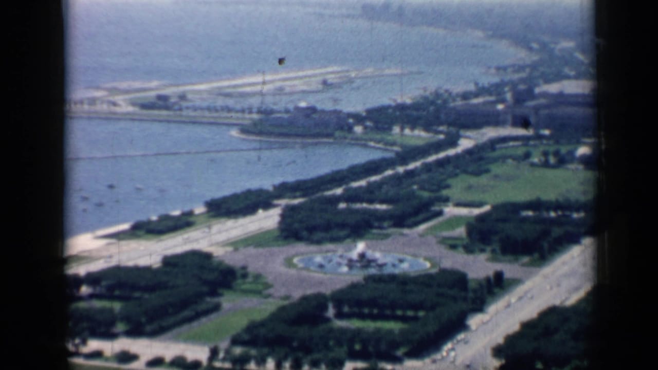 1958 Chicago 8mm frame showing an elevated view over a wooded lakefront park, circular fountain, shoreline, and broad roads.