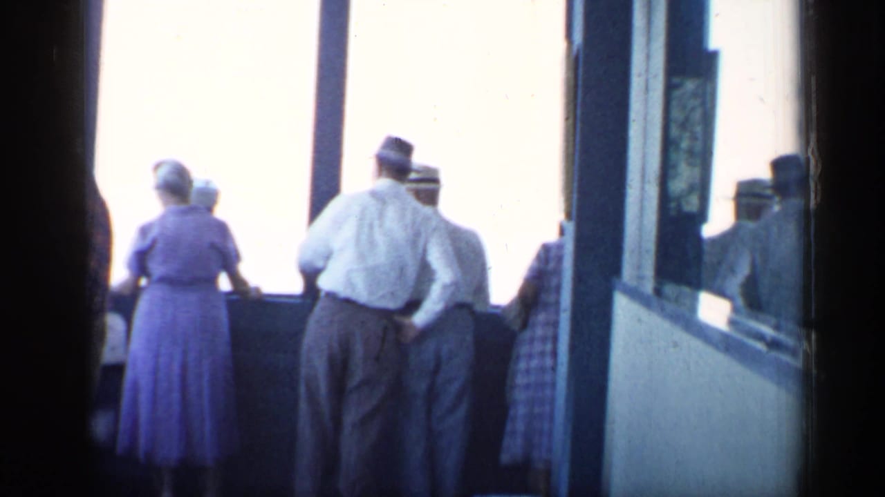 1958 Chicago home-movie frame with adults in period clothing standing at bright windows in an indoor viewing area.