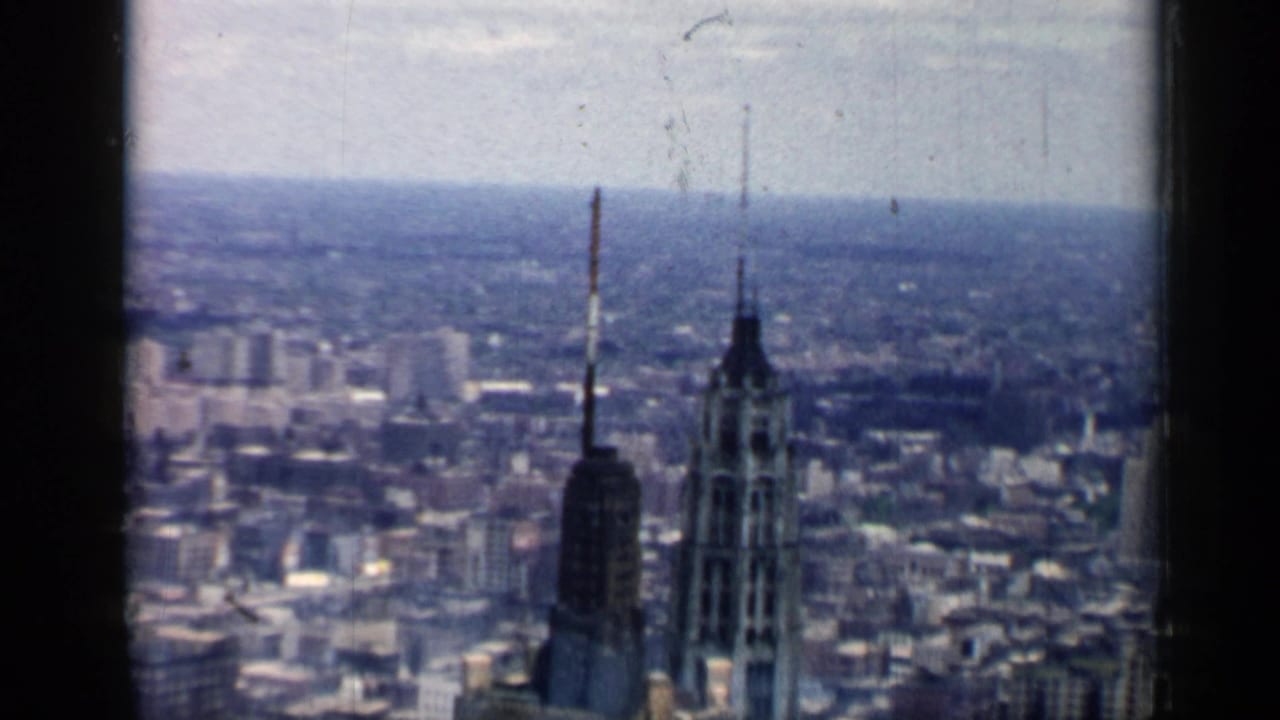 1958 Chicago home-movie frame showing a hazy elevated city panorama with tall towers, antenna masts, and dark 8mm frame edges.