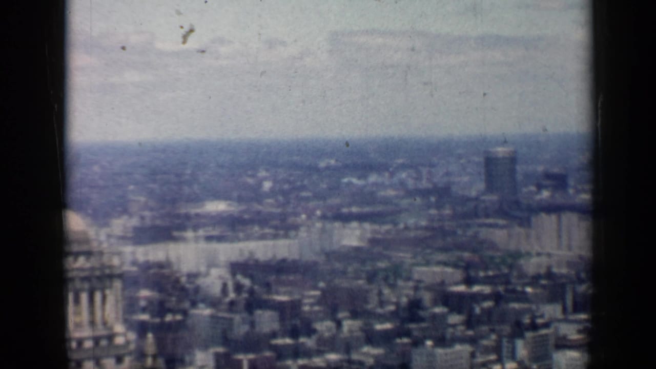 1958 Chicago frame showing a broad skyline from above, with dense buildings, a tall distant tower, and visible film grain.