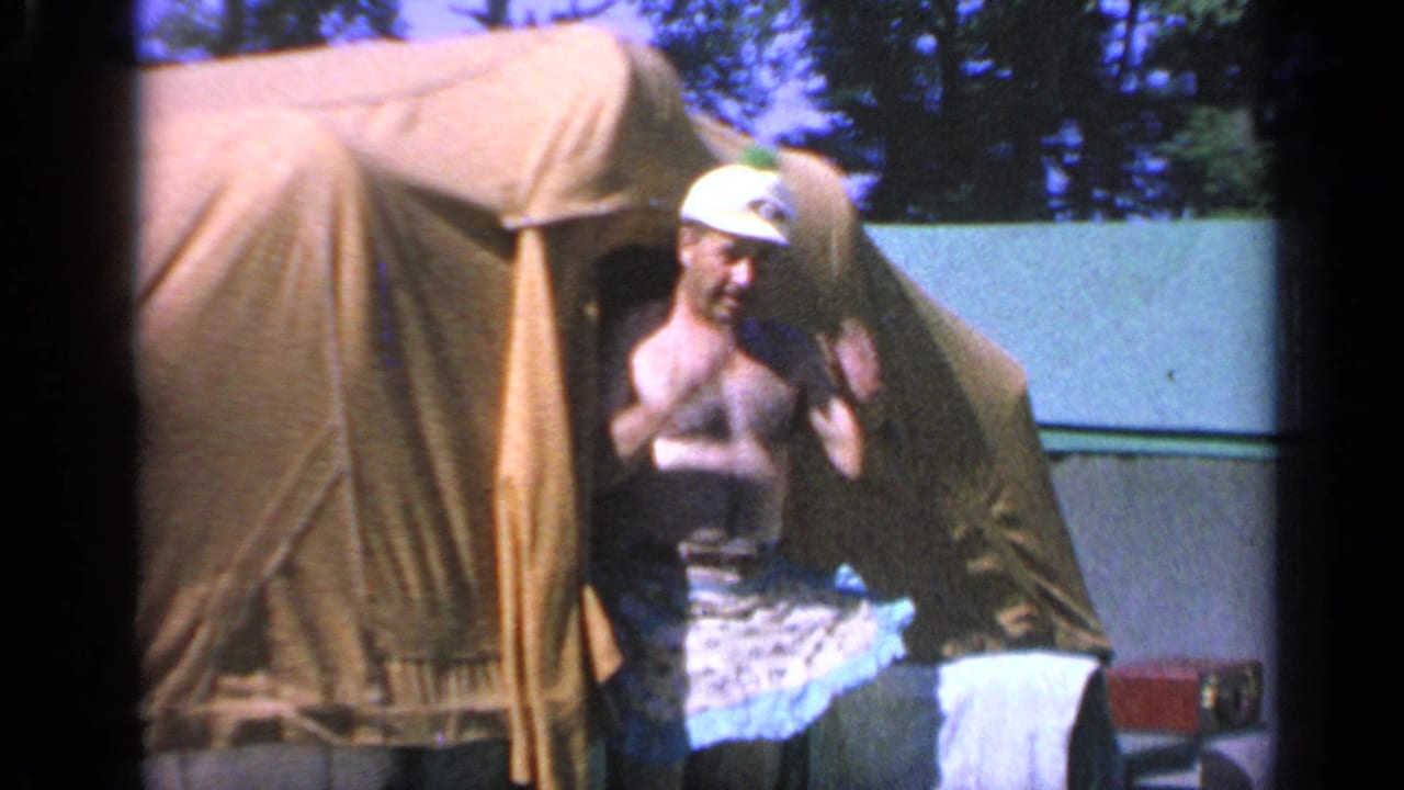 In a 1959 Colorado home-movie frame, a man in a white cap and patterned apron or cloth stands at a tan canvas tent entrance.