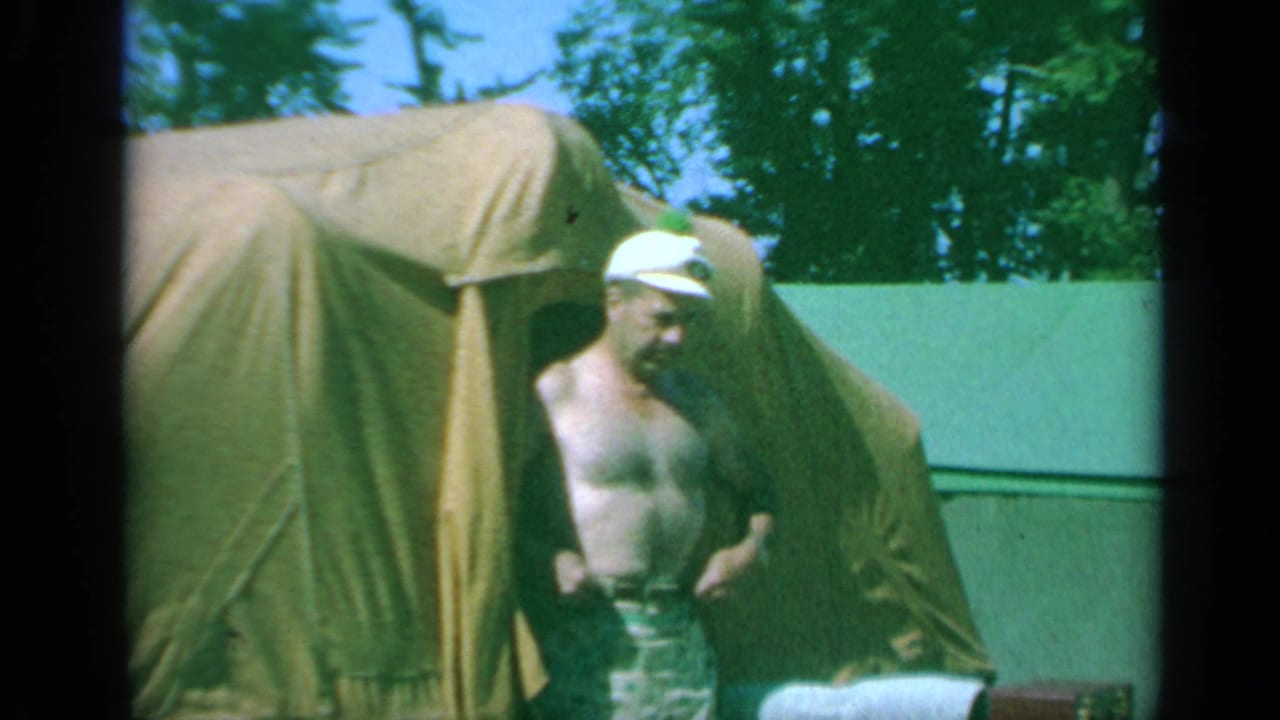 The man stands outside the brown canvas tent, wearing a white cap and patterned shorts, with trees and a green wall behind him.