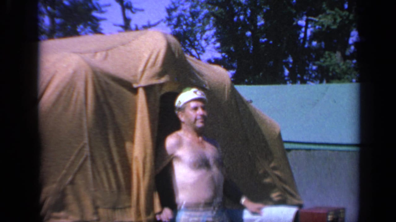 A shirtless man in a white cap stands at the canvas tent entrance, looking aside in a casual campsite moment.
