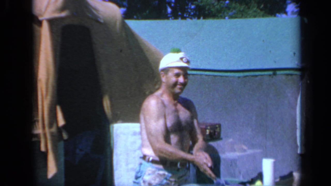 Man in a white cap smiling near a tent and picnic supplies.