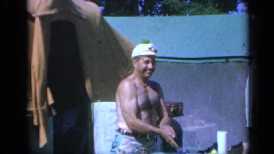 1959 Colorado campsite frame with a man smiling near a tent, cooler, and outdoor food setup.