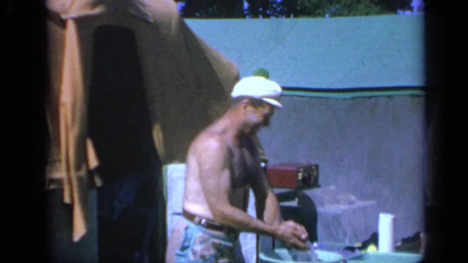 1959 Colorado home-movie frame of a man working with food or supplies beside a tent at a campsite.