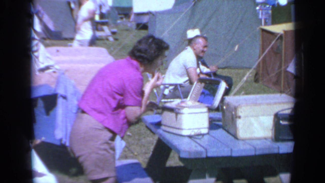 1959 Colorado campsite frame showing a woman at a picnic table with coolers and a seated man nearby.